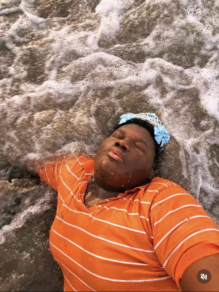 Lex lying in the surf with his eyes closed, wearing a tiara and an orange striped shirt